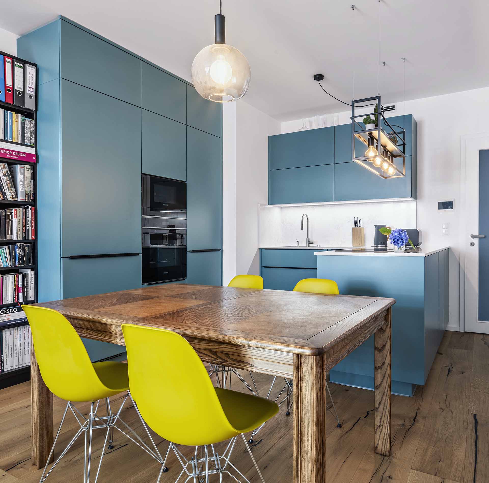Moderní kuchyně s petrolejovými skříňkami, dřevěným jídelním stolem a žlutými židlemi Vitra.